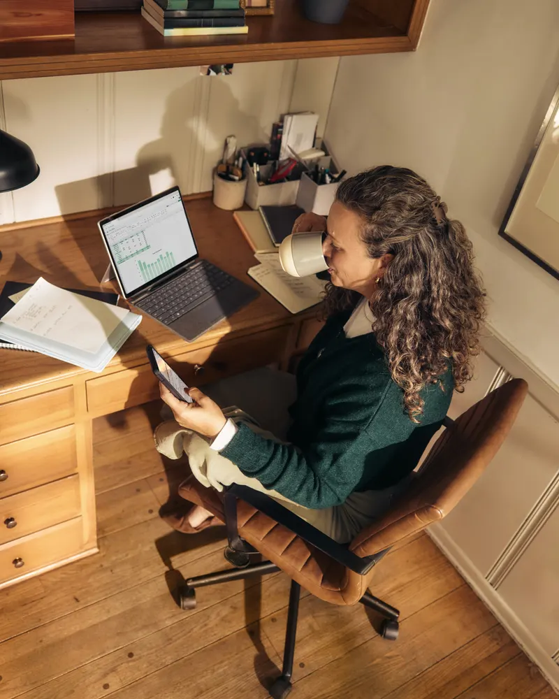 Woman at a home office desk drinking coffee, a spreadsheet open on her laptop